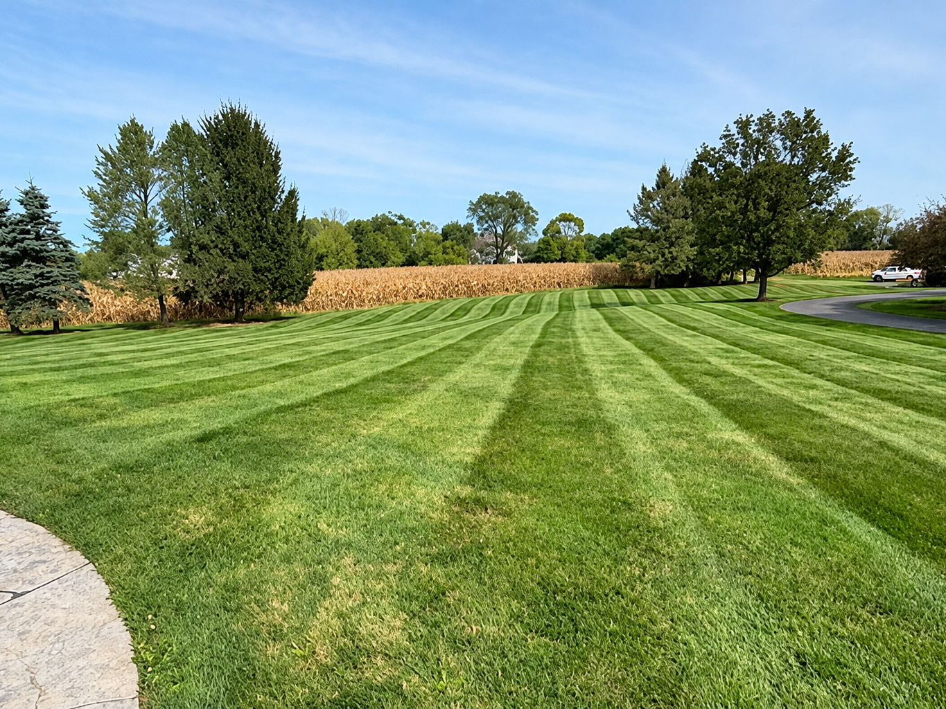 A wide lawn mown in alternating light and dark stripes, bordered by a stone patio and a paved drive
